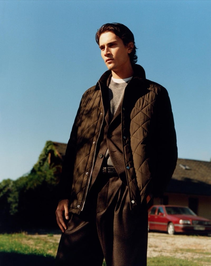 Liam Kelly in a brown quilted jacket over a gray sweater, tailored jacket, and pleated trousers outside a country house