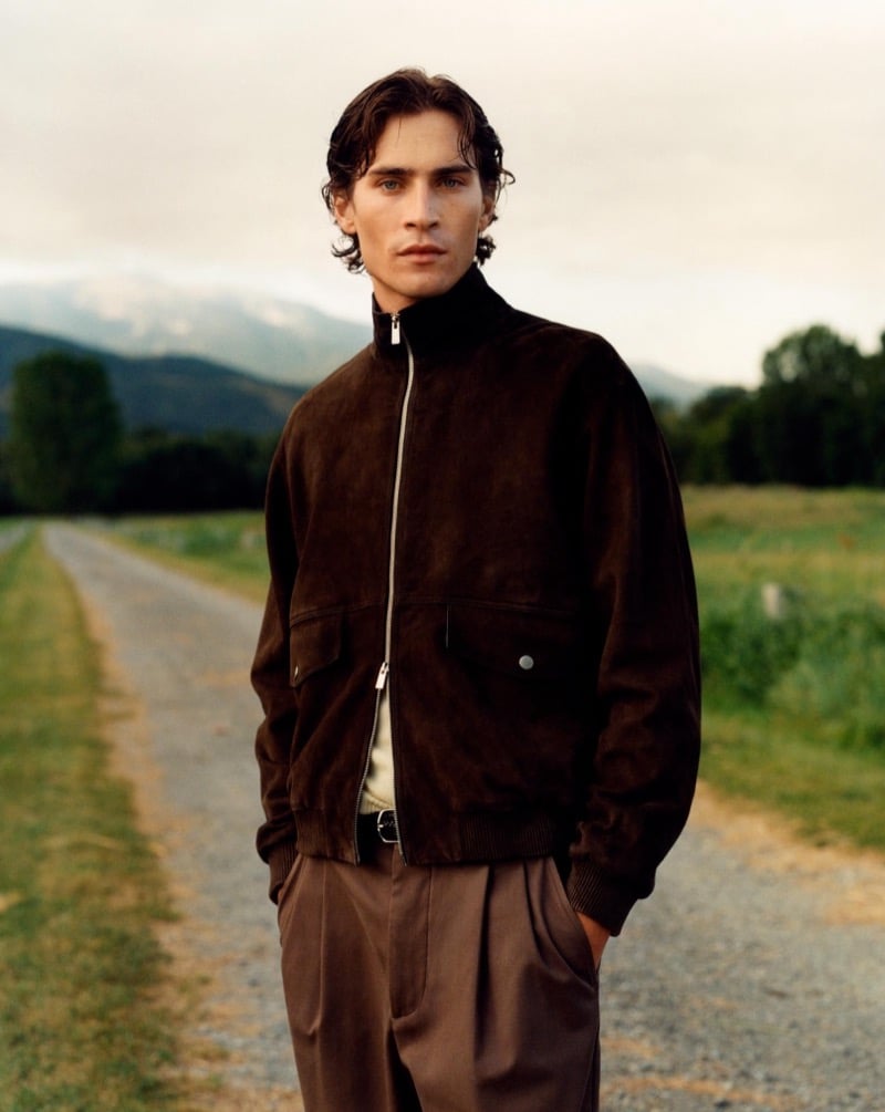 Liam Kelly in a brown suede bomber jacket and pleated trousers on a gravel road with mountains behind him