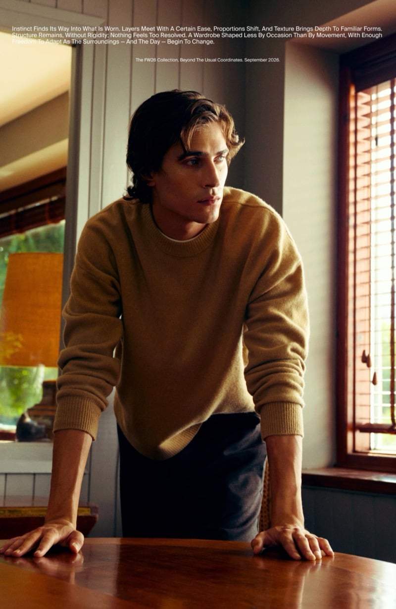 Liam Kelly in a wool crewneck sweater leaning on a wooden table beside a window