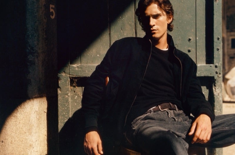 Liam Kelly seated against a weathered wooden door in a zip-up jacket, crewneck sweater, and jeans