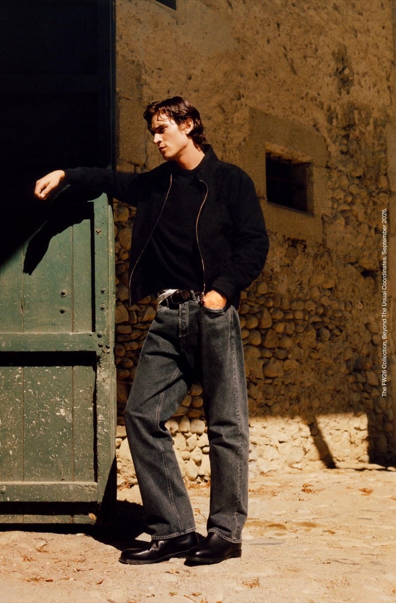 Liam Kelly in a zip-up jacket, belted washed jeans, and leather shoes leaning on a wooden barn door beside a stone wall