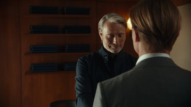 Mads Mikkelsen in a dark shirt smiling toward a younger man seen from behind in a gray suit, shelves of folded knitwear beside them