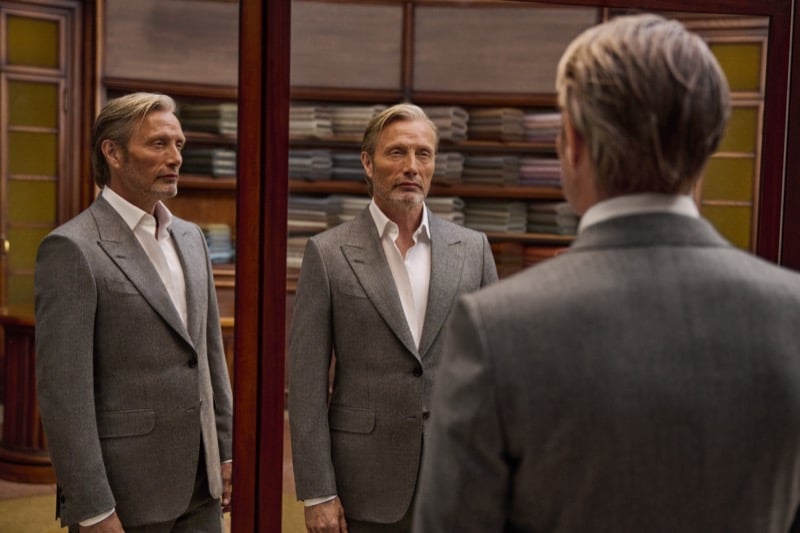 Mads Mikkelsen in a gray tailored jacket and open-collar shirt standing before a three-panel fitting mirror in a wood-paneled Zegna tailoring room lined with stacked bolts of cloth