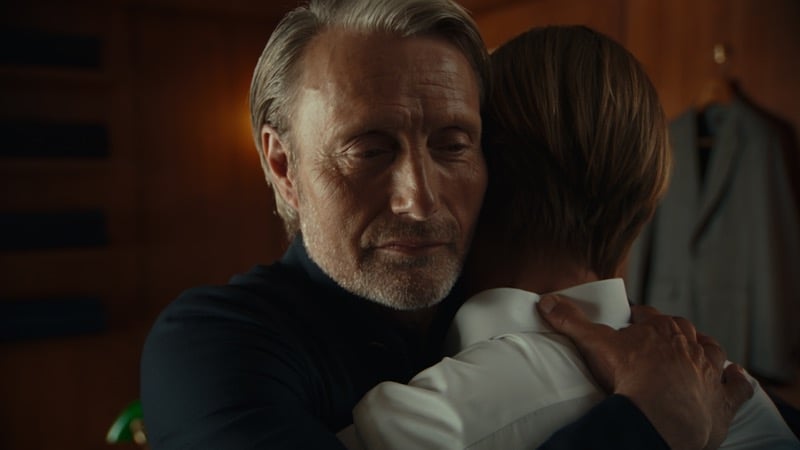 Mads Mikkelsen in a dark knit top embracing a younger blond man in a white shirt, a suit hanging on the wardrobe behind them