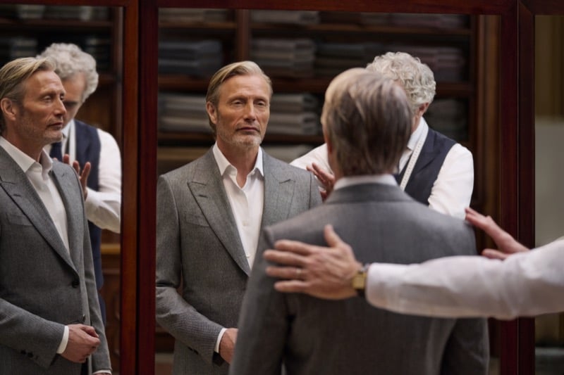 Mads Mikkelsen in a gray suit standing before angled fitting mirrors as a tailor with a measuring tape adjusts the shoulders