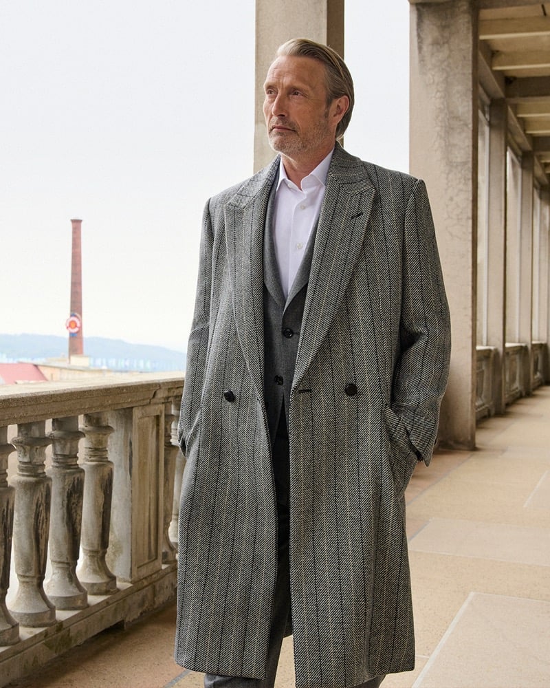 Mads Mikkelsen on a stone colonnaded terrace in a gray herringbone double-breasted overcoat over a gray suit, hands in his coat pockets, an industrial chimney in the distance