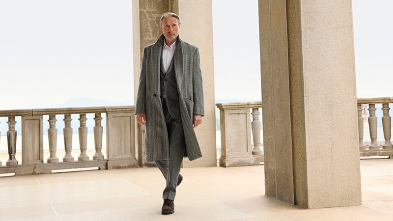 Mads Mikkelsen walking along a stone terrace colonnade in a gray herringbone overcoat over a gray suit, water and hills behind him