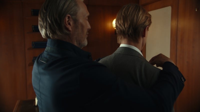 Mads Mikkelsen adjusting the collar of a gray jacket on a younger man facing a mirror in the wood-paneled fitting room