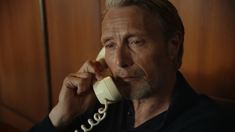 Close-up of Mads Mikkelsen in a dark shirt holding a cream vintage telephone to his ear