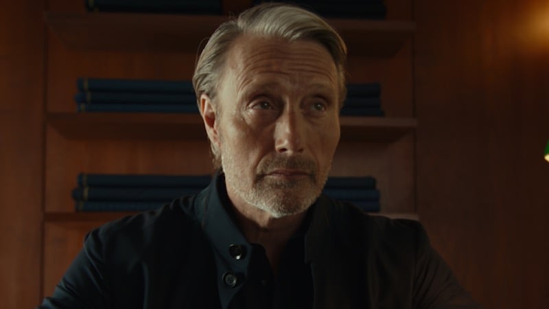 Close-up portrait of Mads Mikkelsen in a dark shirt seated against shelves stacked with folded cloth