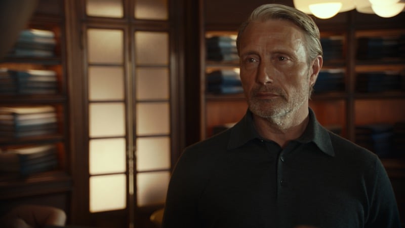 Mads Mikkelsen in a dark knit polo standing in a warmly lit wood-paneled Zegna salon lined with folded sweaters