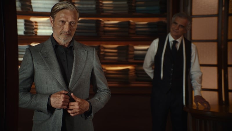 Mads Mikkelsen buttoning a gray suit jacket over a dark shirt as a tailor in a waistcoat watches from a doorway
