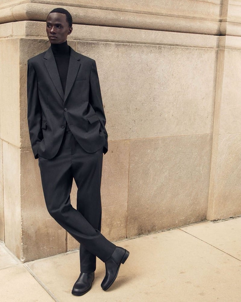 Malick Bodian leans against a stone wall with his hands in his pockets in a blazer over a turtleneck with trousers and leather ankle boots, for COS's fall 2026 campaign
