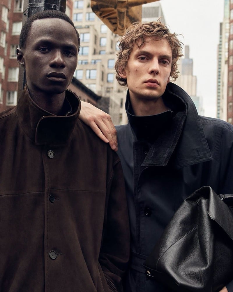 Malick Bodian in a brown suede jacket and Leon Dame in a trench with a leather bag stand shoulder to shoulder, Leon's hand on Malick's shoulder, for COS's fall 2026 campaign