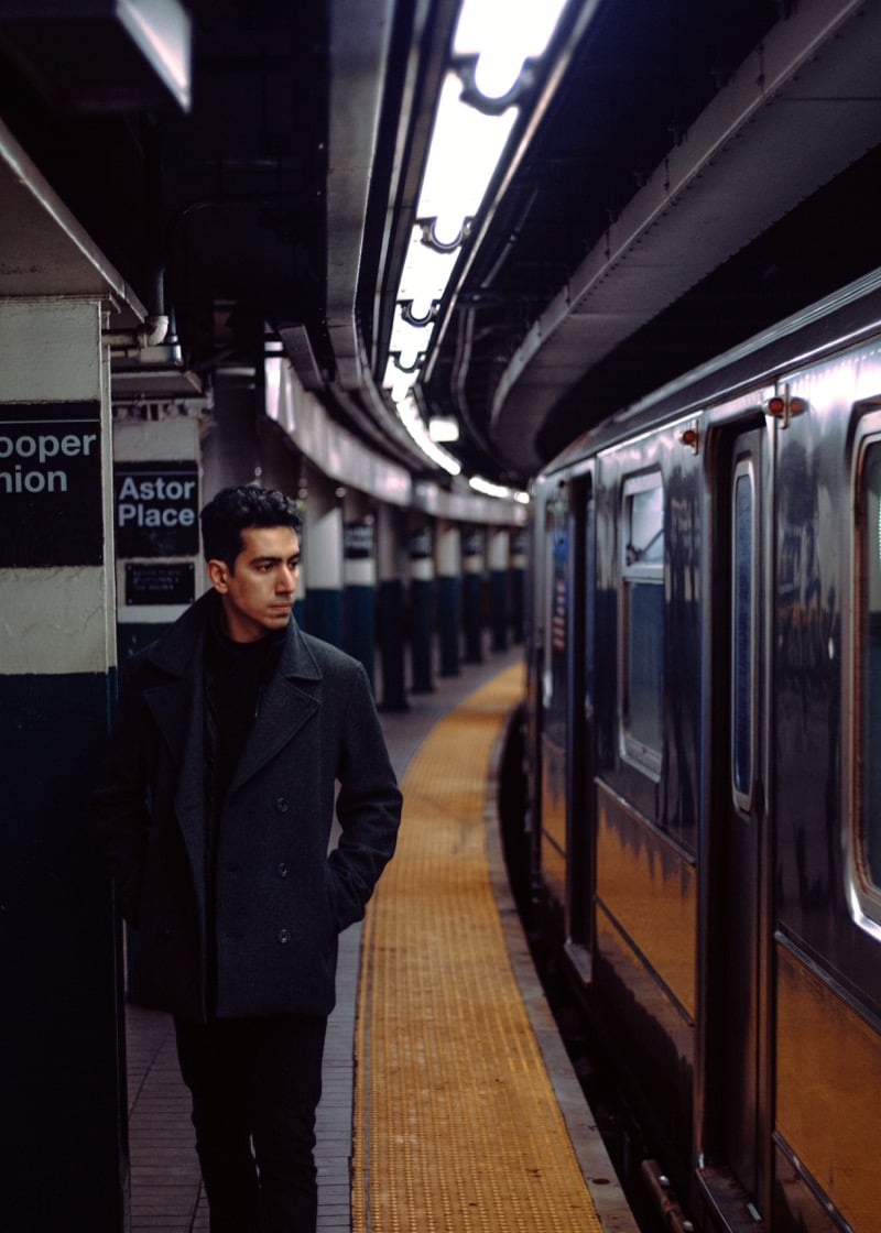 A man in a charcoal peacoat stands against a pillar on a subway platform as a train waits alongside