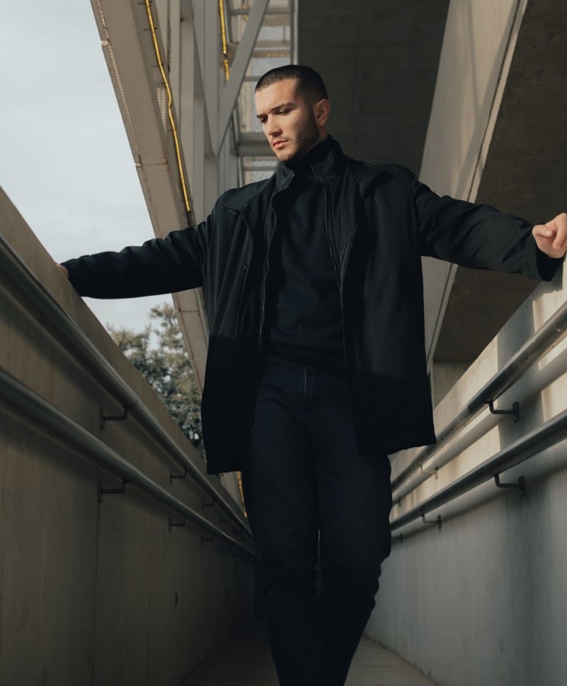 A man in a black jacket, black turtleneck, and dark jeans stands on a concrete walkway in flat daylight
