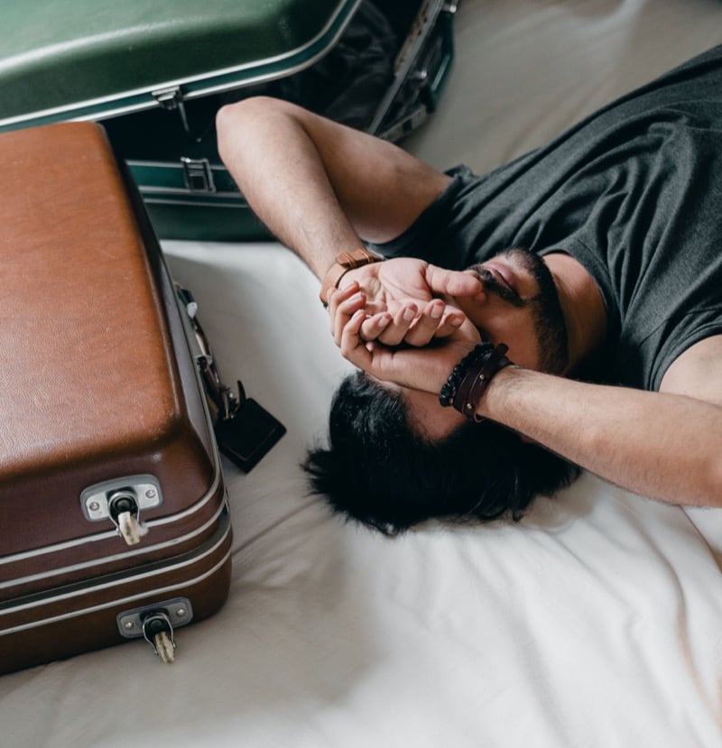 A man lies on a bed with his hands over his face, surrounded by open vintage suitcases