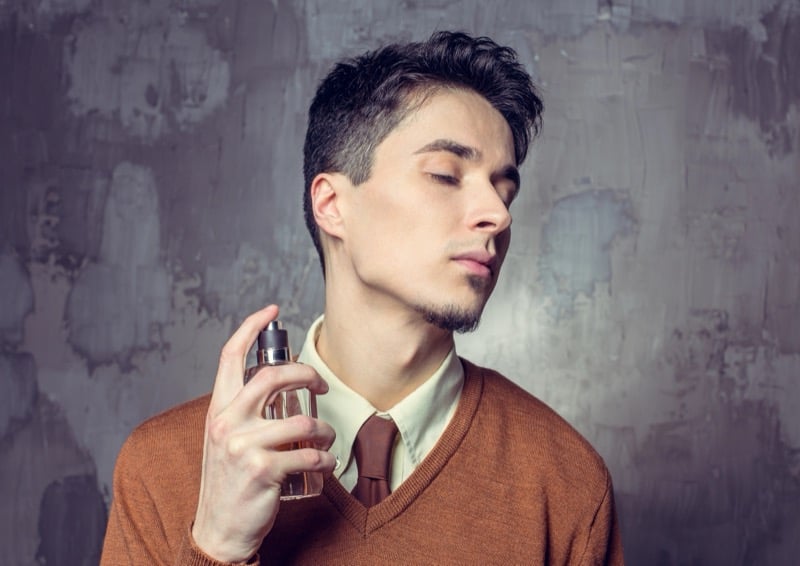 Man spraying cologne toward his neck in a brown V-neck sweater and tie against a textured gray wall