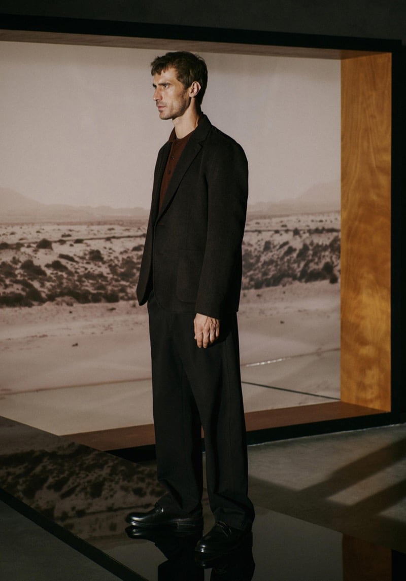 Profile view of Clément Chabernaud standing in front of a large desert landscape print, wearing a dark brown blazer over a brown knit polo with matching trousers and black loafers