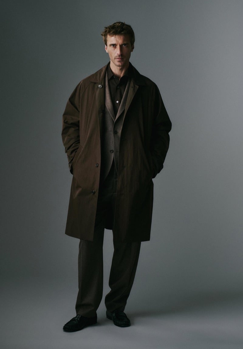 Clément Chabernaud stands in a studio in a long brown car coat worn open over a brown suit and a button-up shirt, hands in his coat pockets, with dark loafers