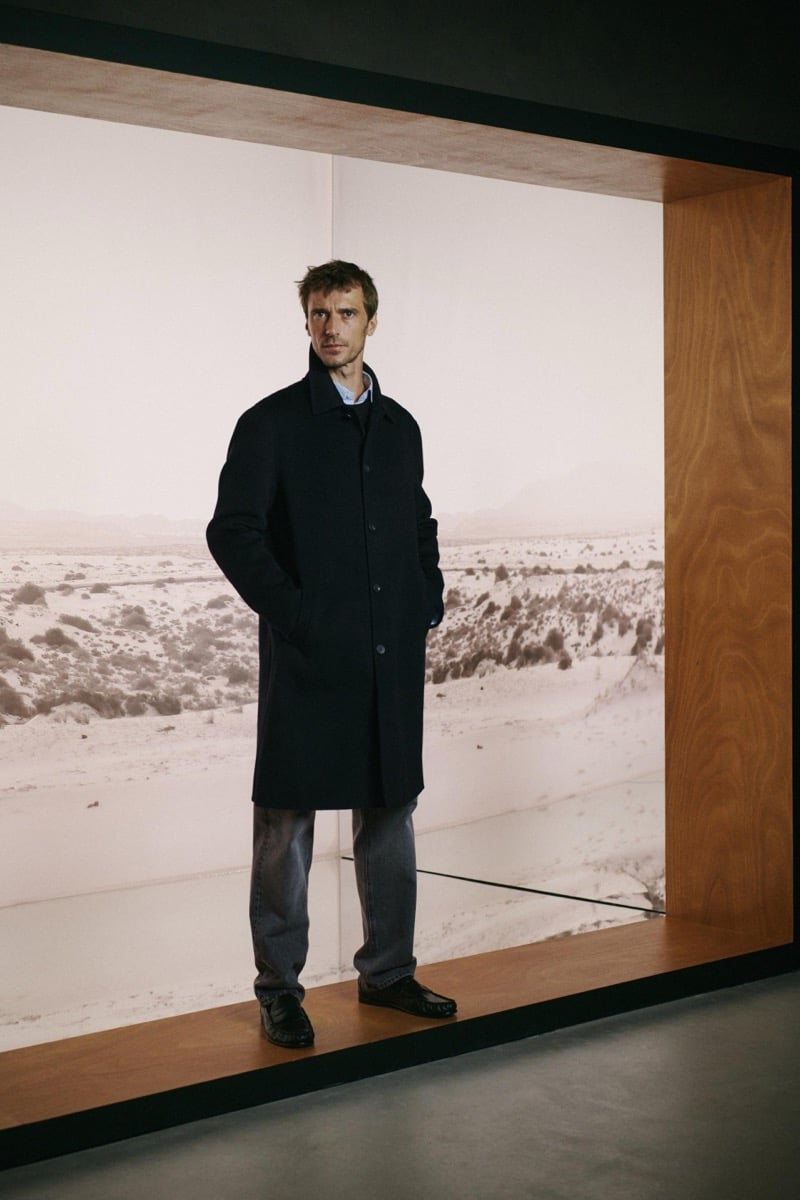 Clément Chabernaud stands in front of a lit desert landscape display in a dark knee-length car coat over a light blue shirt, with faded gray jeans and black moccasins