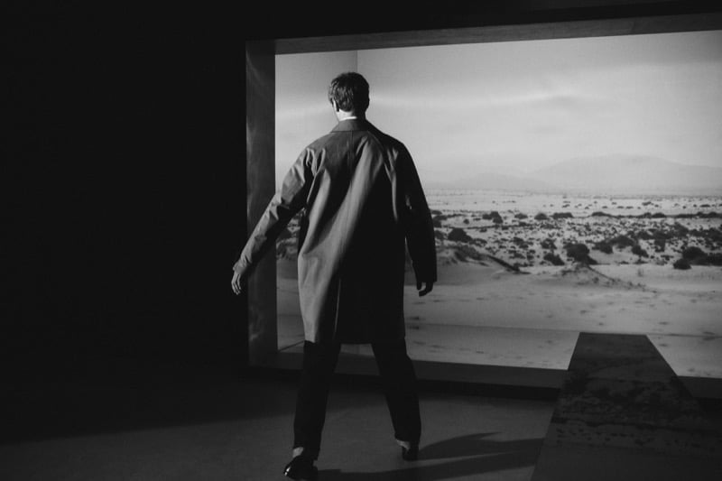 Black-and-white photo of Clément Chabernaud seen from behind in a knee-length car coat, walking toward a large illuminated desert landscape print in a dark gallery space