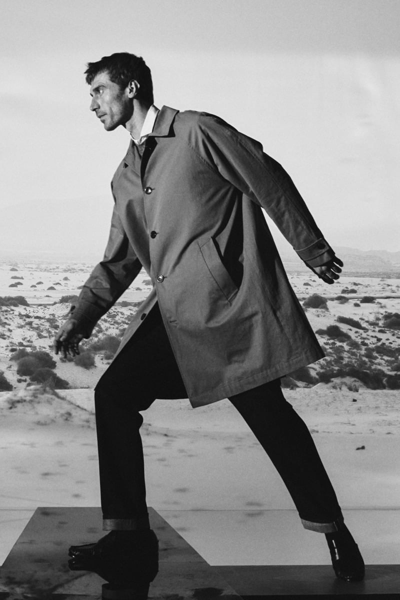 Black-and-white photo of Clément Chabernaud mid-stride on a raised platform in front of a desert backdrop, wearing an open knee-length car coat over a shirt, cuffed trousers, leather gloves, and glossy loafers