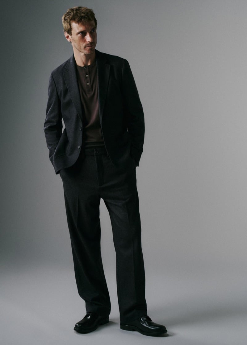Clément Chabernaud in a charcoal soft-tailored blazer over a brown henley knit and dark pleated trousers, hands in pockets, with black loafers