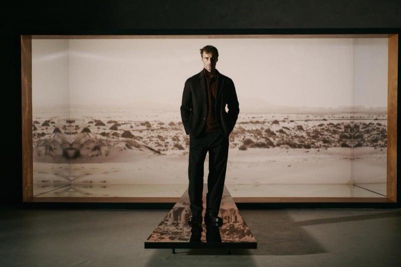 Clément Chabernaud stands on a low platform in a gallery space in front of a large desert landscape photograph, wearing a charcoal wool jacket over a brown knit with dark tapered trousers and black boots