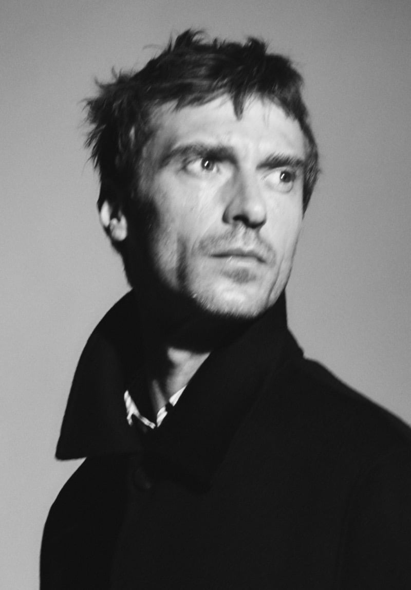Grainy black-and-white portrait of Clément Chabernaud with windswept hair, wearing a dark coat with the collar up over a striped shirt, looking off to the side