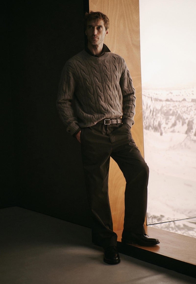 Clément Chabernaud leans against a wood panel beside an illuminated snowy landscape image, wearing a gray cable-knit sweater over a dark collared shirt with dark wide trousers, a leather belt, and black derby shoes