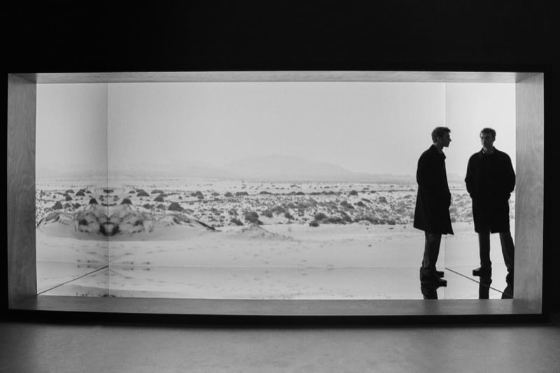 Black-and-white wide shot of Clément Chabernaud in a dark coat standing at the edge of a large illuminated desert landscape installation, his reflection mirrored in the glass beside him