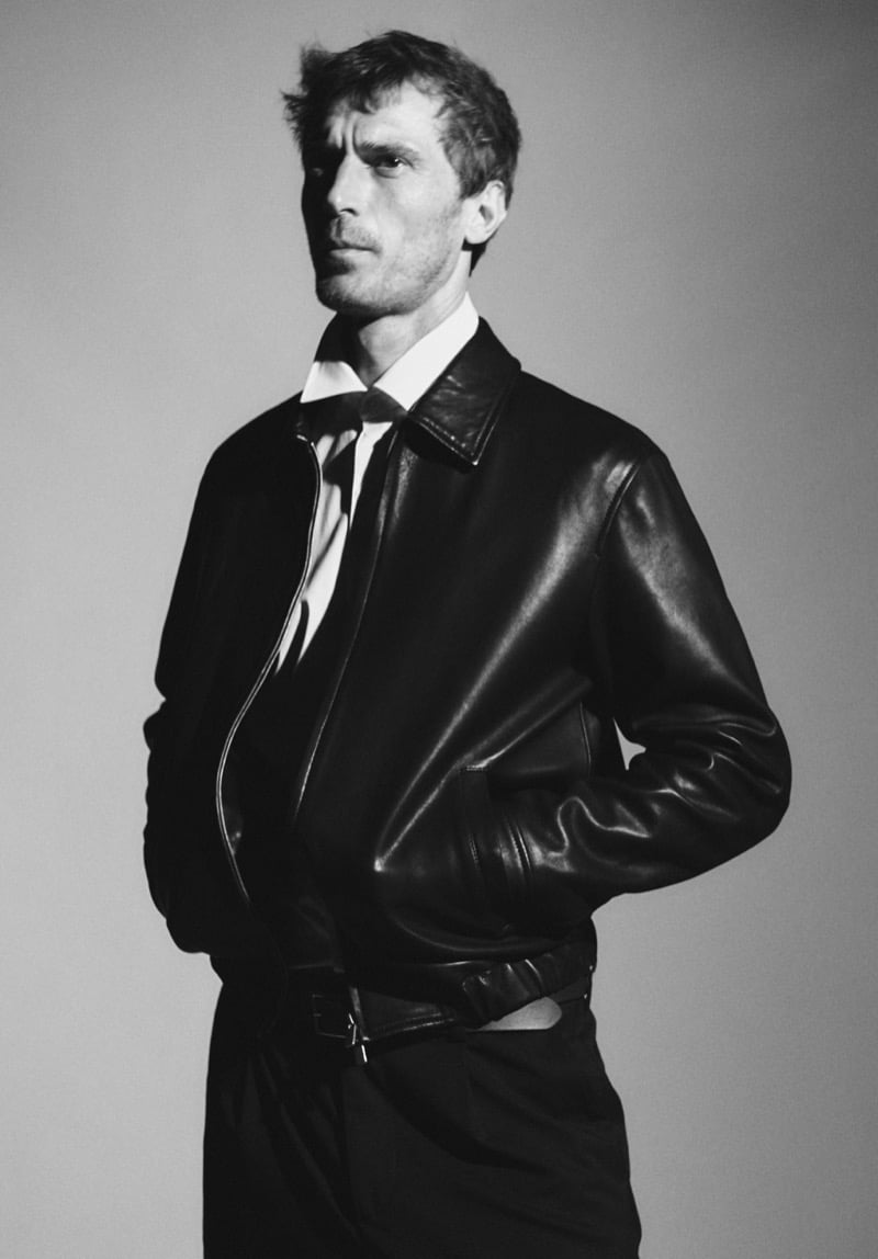 Black-and-white portrait of Clément Chabernaud in a black leather jacket with a shirt collar and loosened tie, hands in his pockets, against a plain backdrop