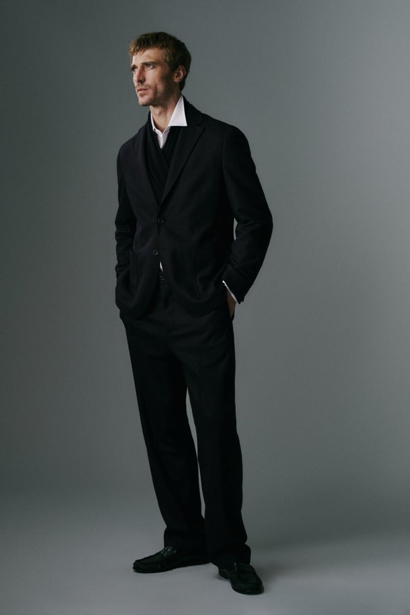 Full-length photo of Clément Chabernaud in a soft dark tailored jacket and matching wide trousers over a scarf and white shirt, hands in pockets, wearing black tasseled loafers
