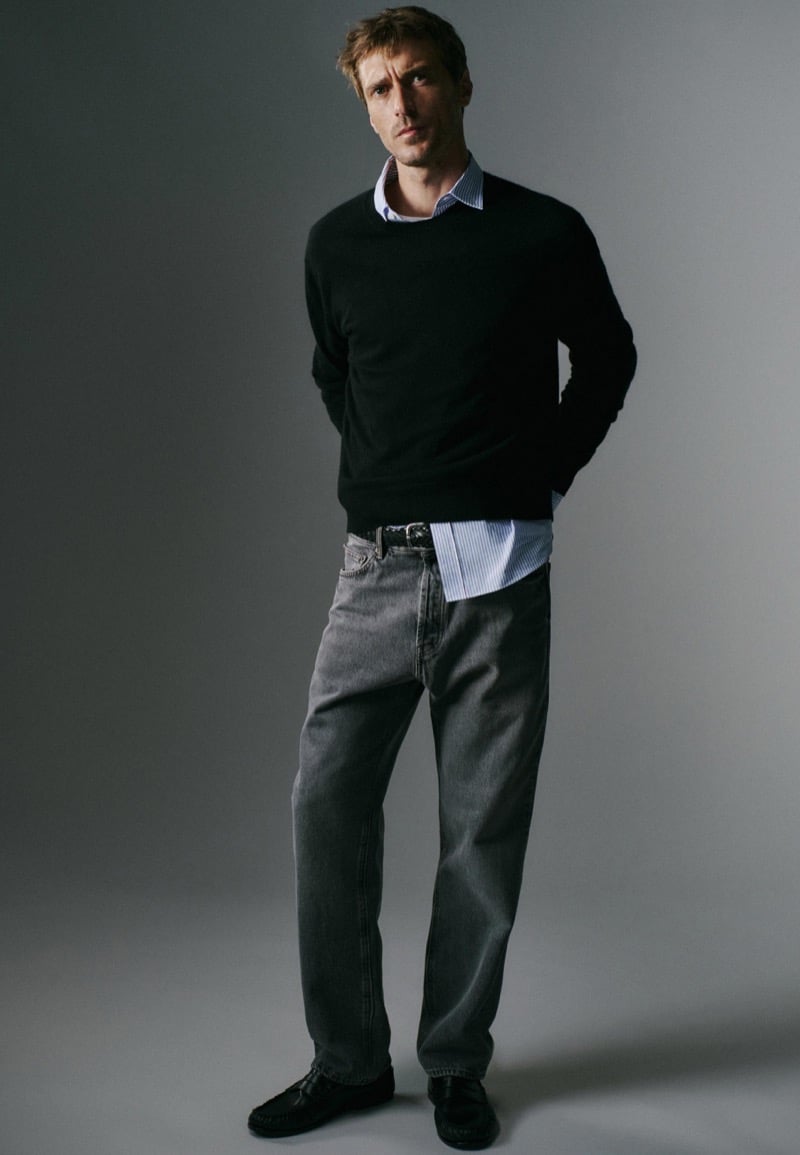 Clément Chabernaud stands with hands in his pockets wearing a dark crew-neck sweater over a striped shirt, washed gray straight-leg jeans, a studded belt, and black moccasins