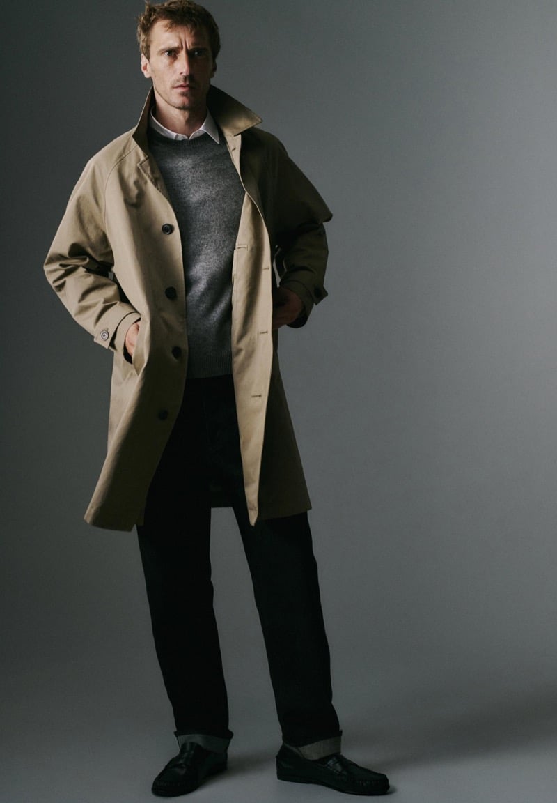 Clément Chabernaud stands in a studio in an open tan car coat with a raised collar over a gray crew-neck sweater and white shirt, with dark cuffed trousers and black tasseled loafers