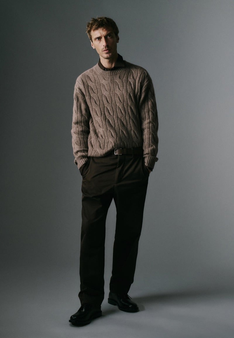 Clément Chabernaud stands in a studio wearing a taupe cable-knit sweater over a dark collared shirt, tucked into dark brown trousers with a leather belt and black derby shoes