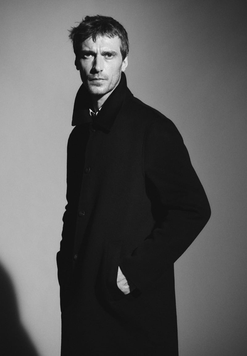 Black-and-white three-quarter portrait of Clément Chabernaud in a single-breasted wool overcoat with a large collar turned up, one hand in his pocket, a striped shirt visible at the neck