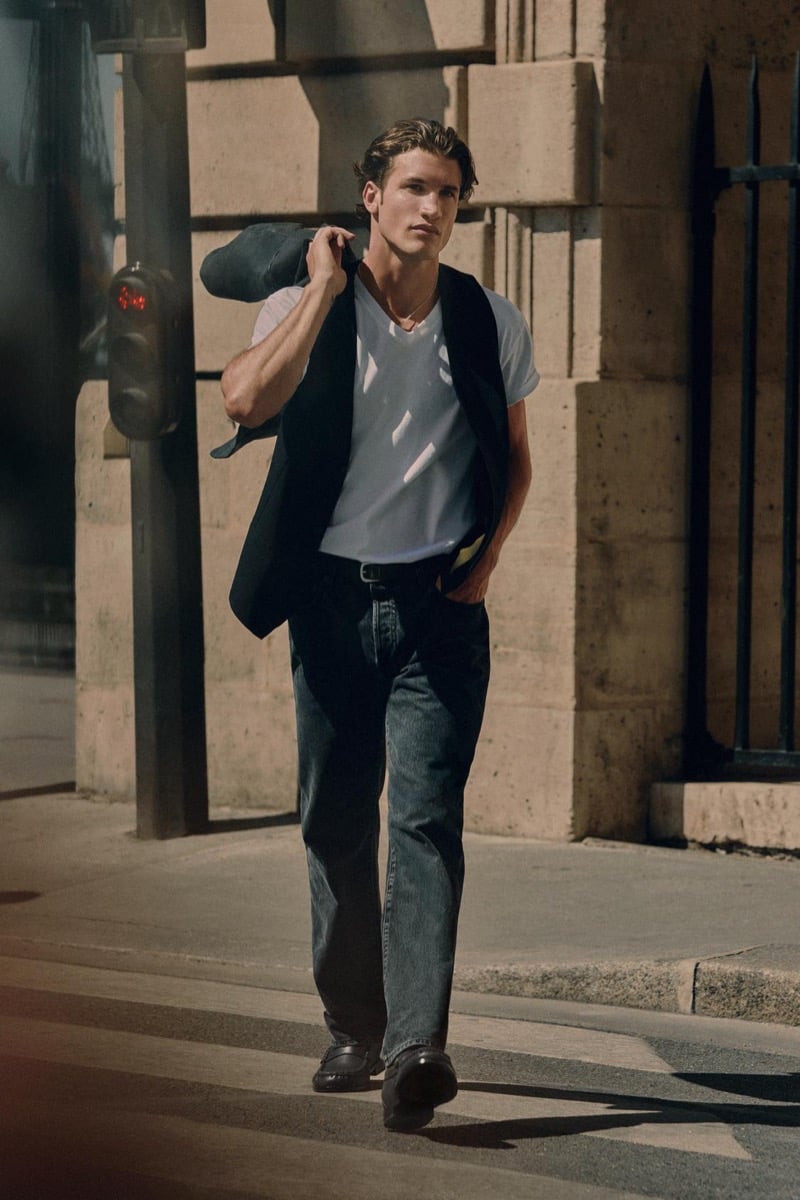 Max Knott crosses the street with a blazer slung over his shoulder, wearing a white v-neck tee and jeans for Zara's 90's Archive