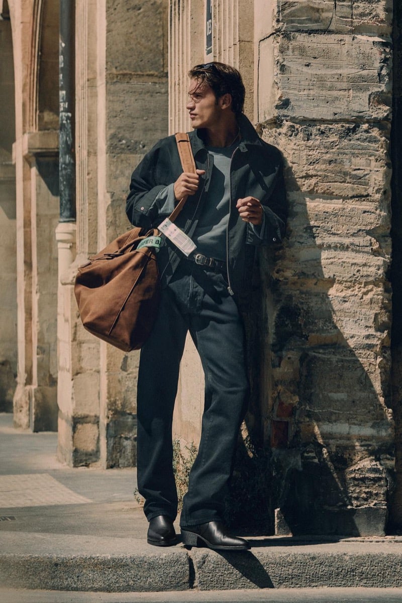 Max Knott leans against a building in a zip jacket with a duffel bag and straight jeans for Zara's 90's Archive