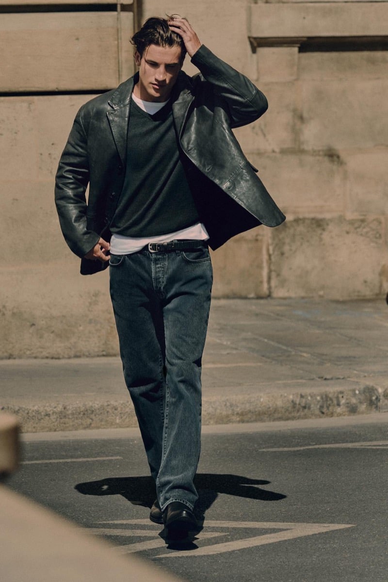 Max Knott walks in a leather-look jacket, sweater, and white tee with straight jeans for Zara's 90's Archive