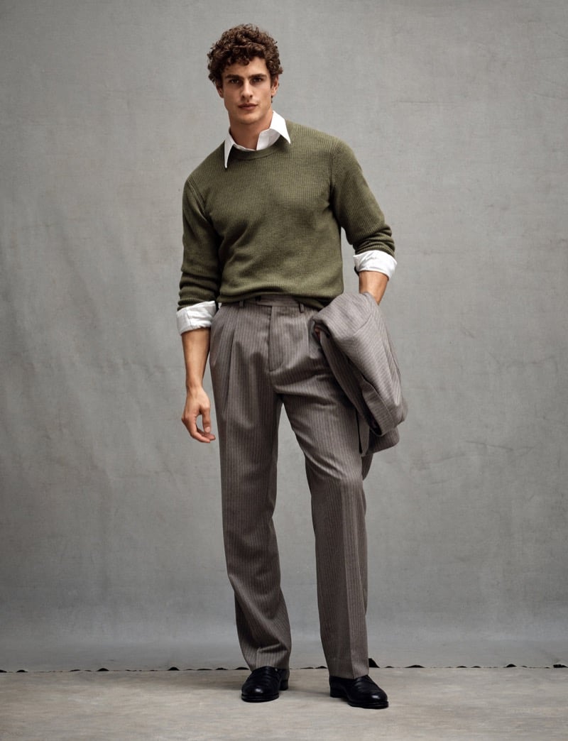 Max Mersmann stands in an olive green sweater layered over a collared shirt with pinstriped pleated trousers, a jacket in hand, and black loafers, for Todd Snyder's Essential Pieces