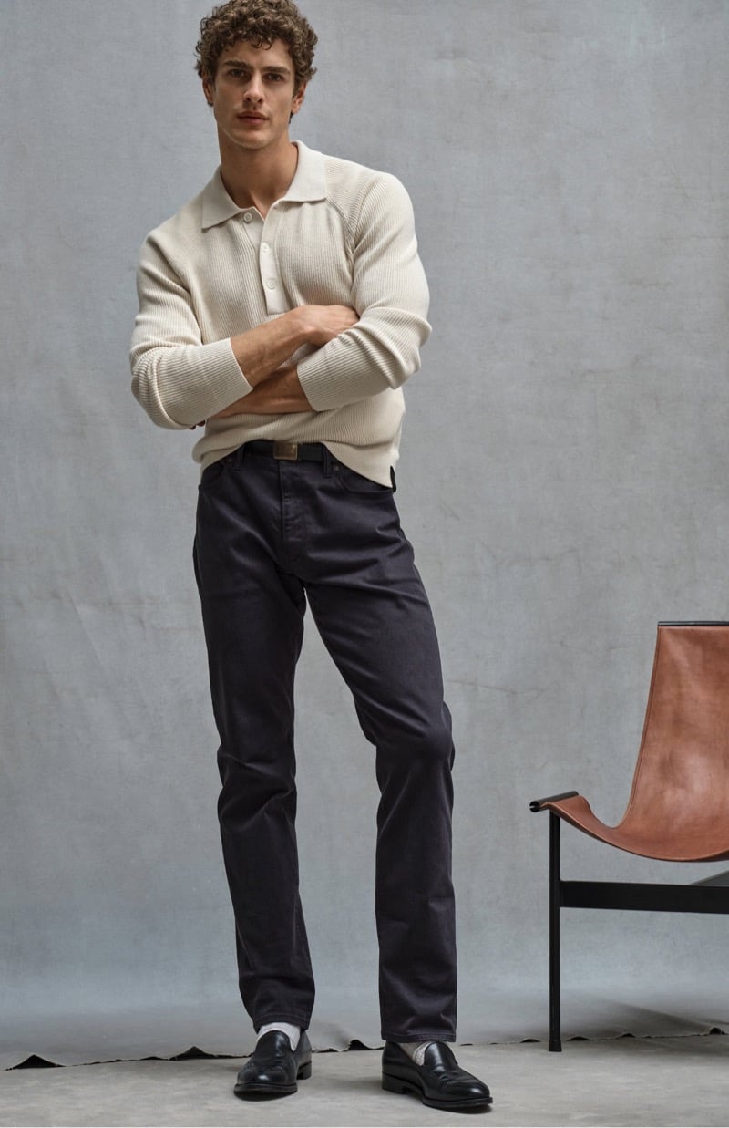 Max Mersmann crosses his arms in a ribbed knit polo with slim 5-pocket chinos and black loafers beside a leather sling chair, for Todd Snyder's Essential Pieces