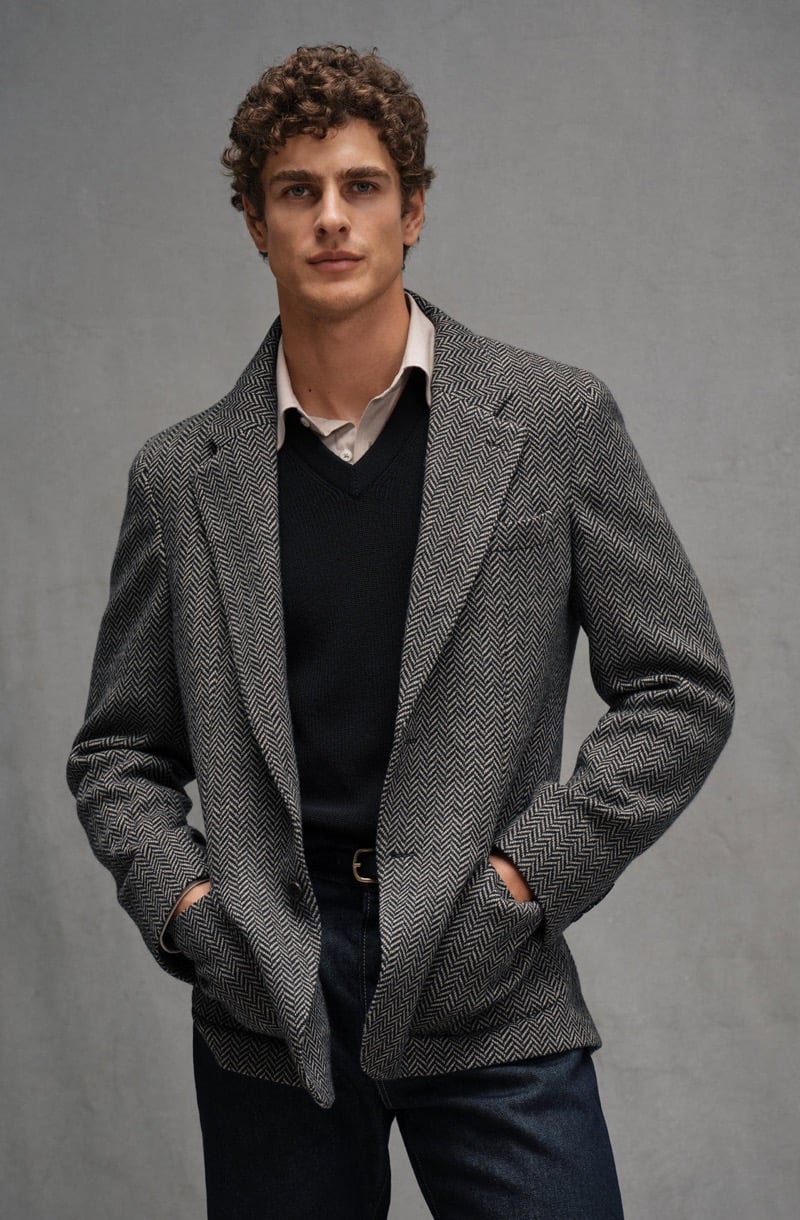 Max Mersmann stands with his hands in the pockets of a herringbone sport coat layered over a V-neck sweater and collared shirt with dark jeans, for Todd Snyder's Essential Pieces