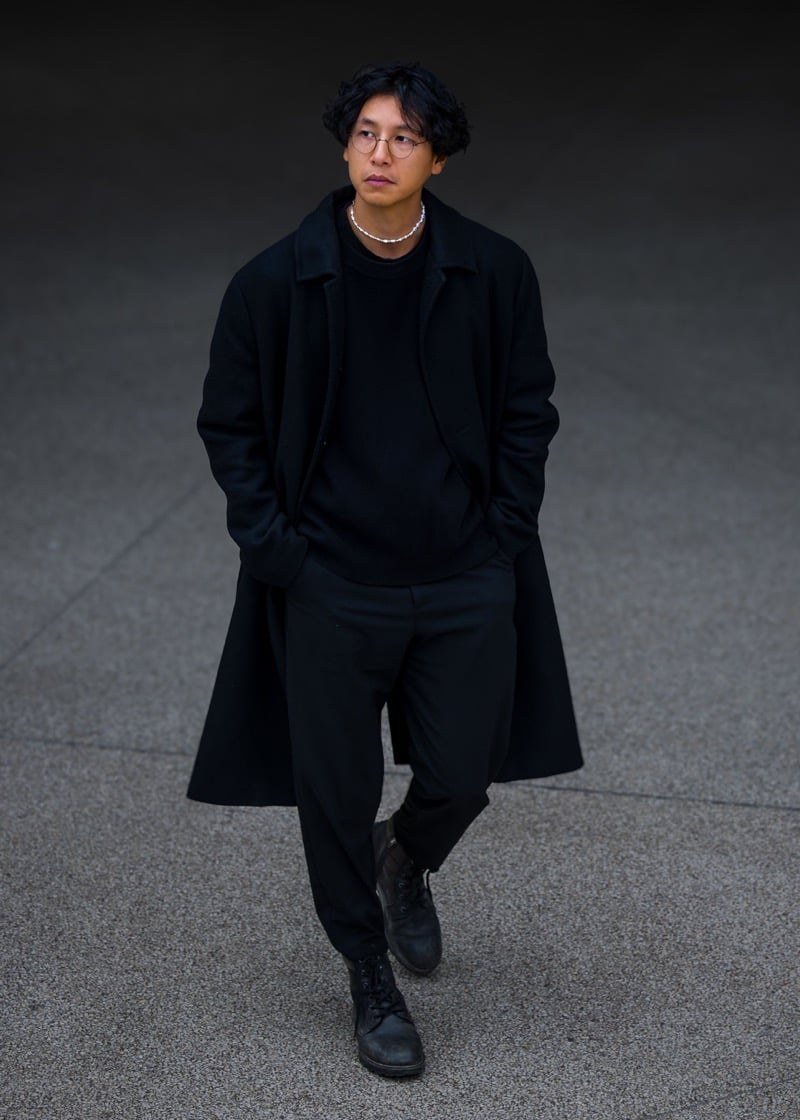A man dressed entirely in black with round glasses, a silver beaded necklace, an unstructured coat, and worn lace-up boots