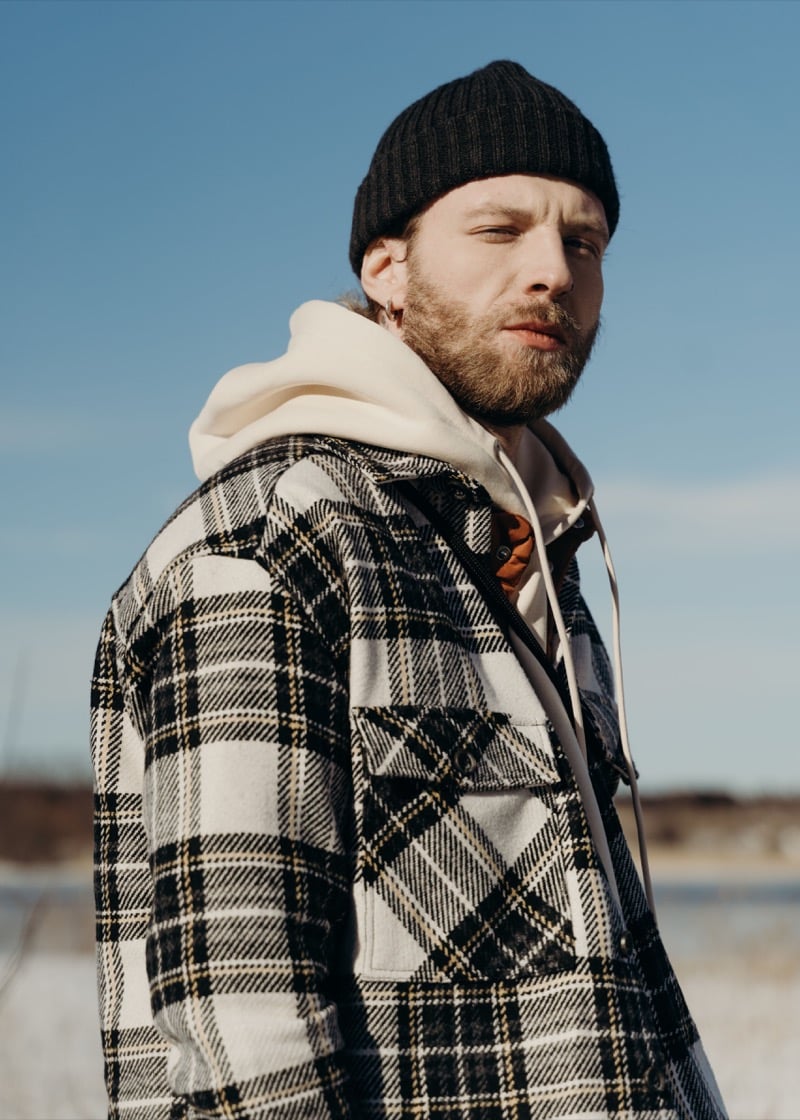 A man in a black ribbed beanie, cream hoodie, and black-and-white plaid wool overshirt outdoors in winter light