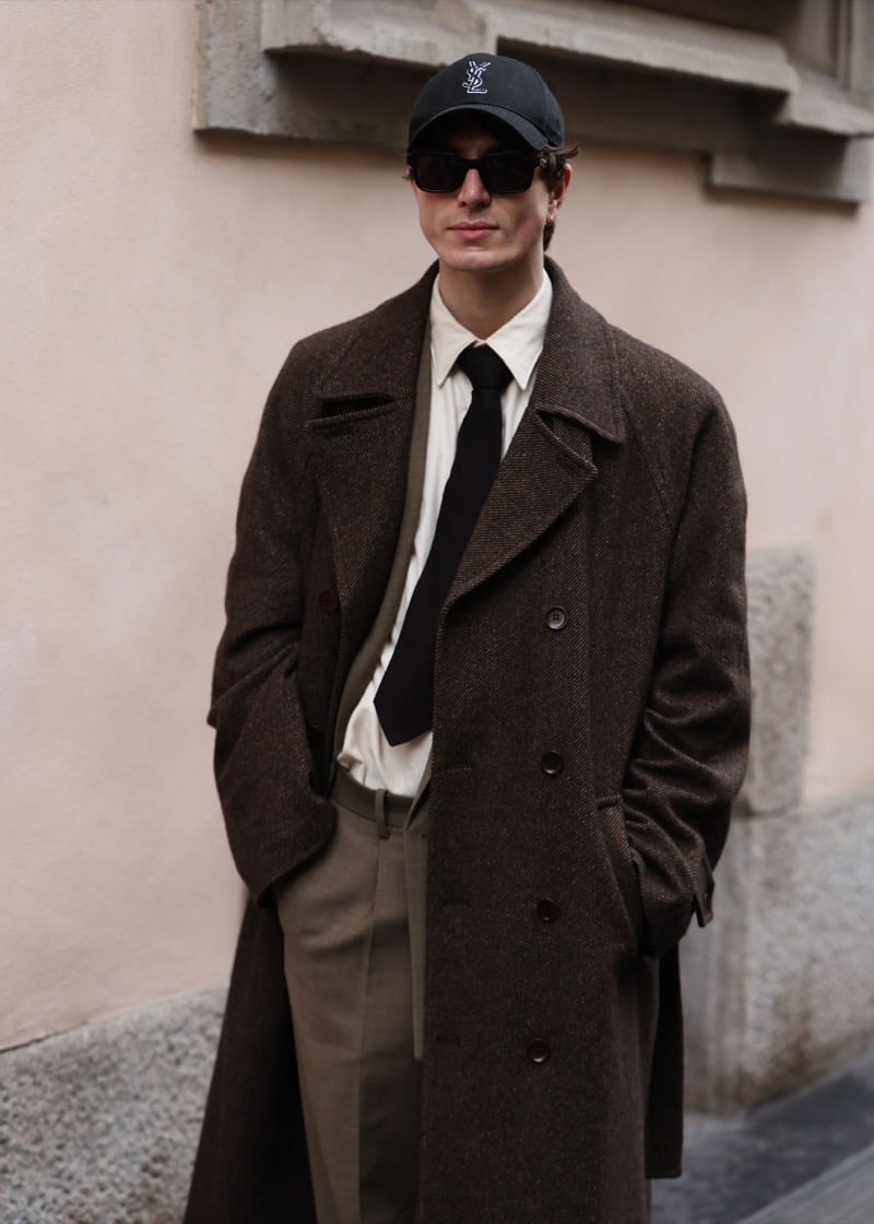 A man in a black cap and sunglasses wearing a brown herringbone overcoat, cream shirt, black tie, and olive trousers against a pale wall