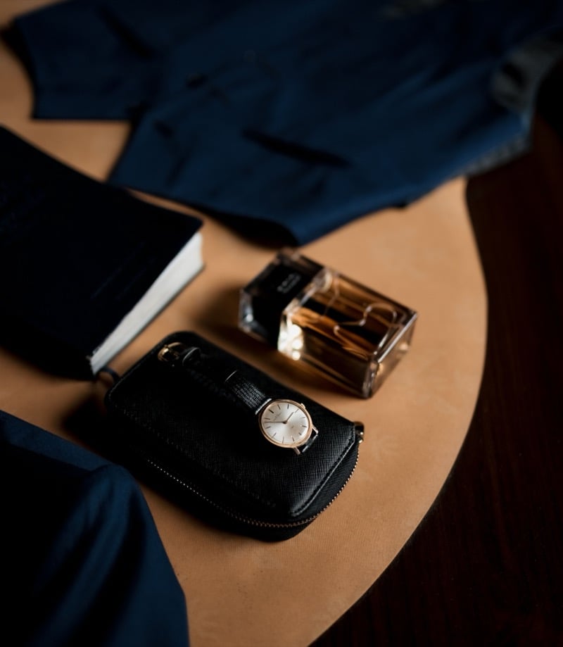 Fragrance bottle beside a gold-tone watch, black leather zip case, notebook and dark suiting on a wooden table