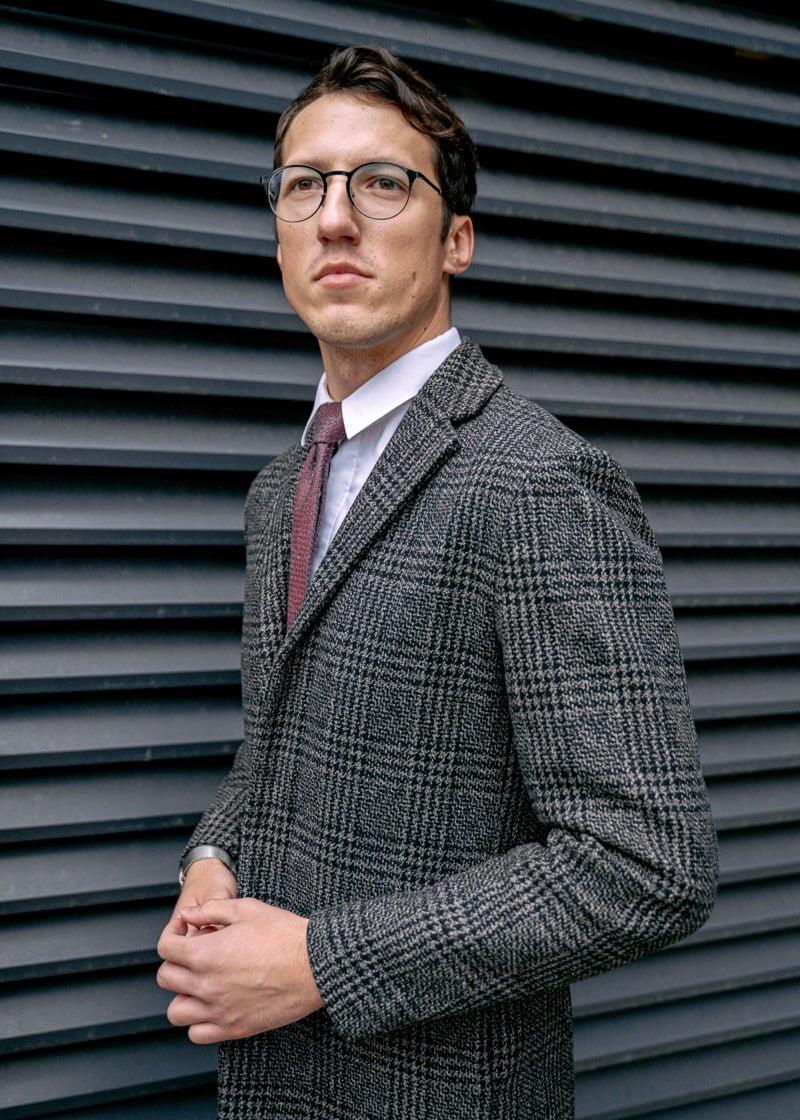 A man in round glasses and a gray Prince of Wales check overcoat with a burgundy knit tie and a wristwatch