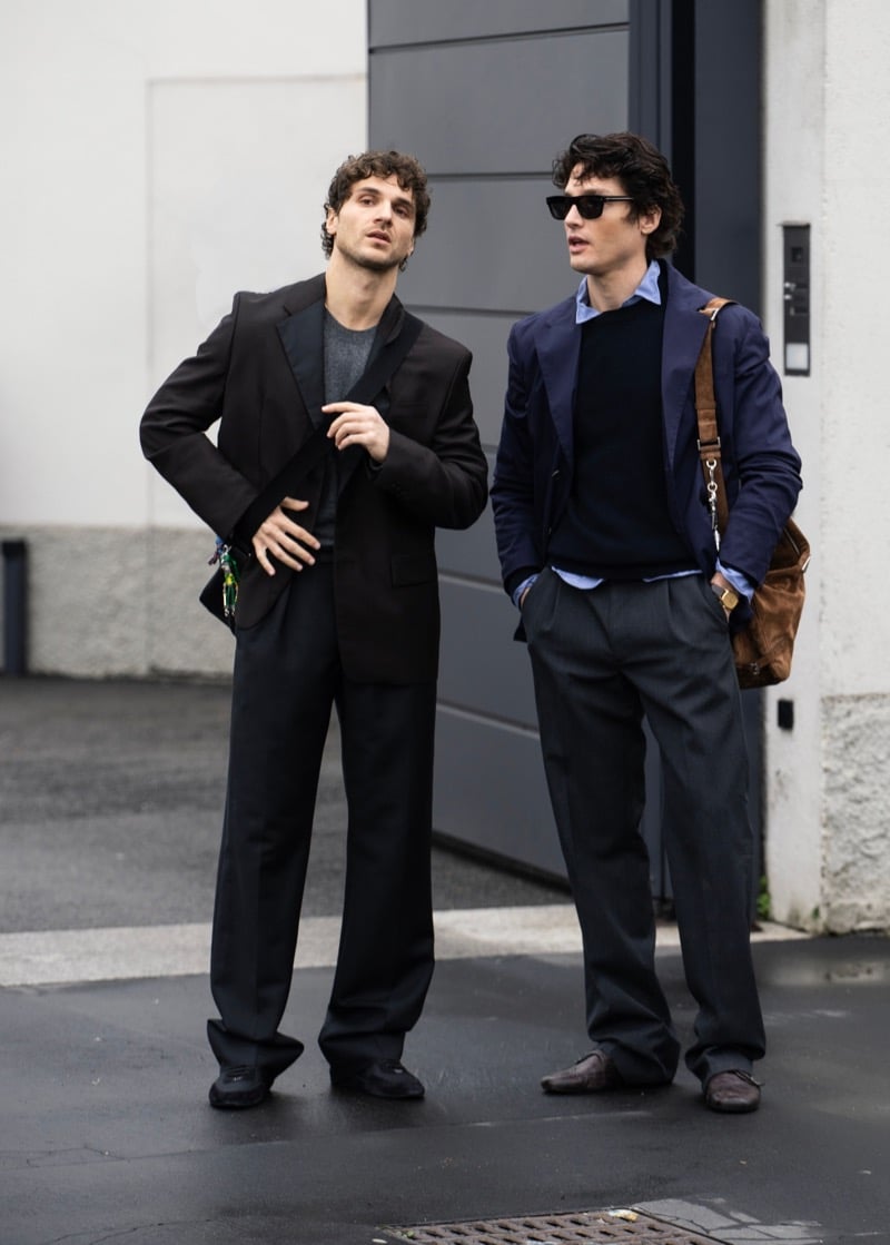 Two men in tailoring on a street, one with a brown suede crossbody bag and black sunglasses, the other in a black suit